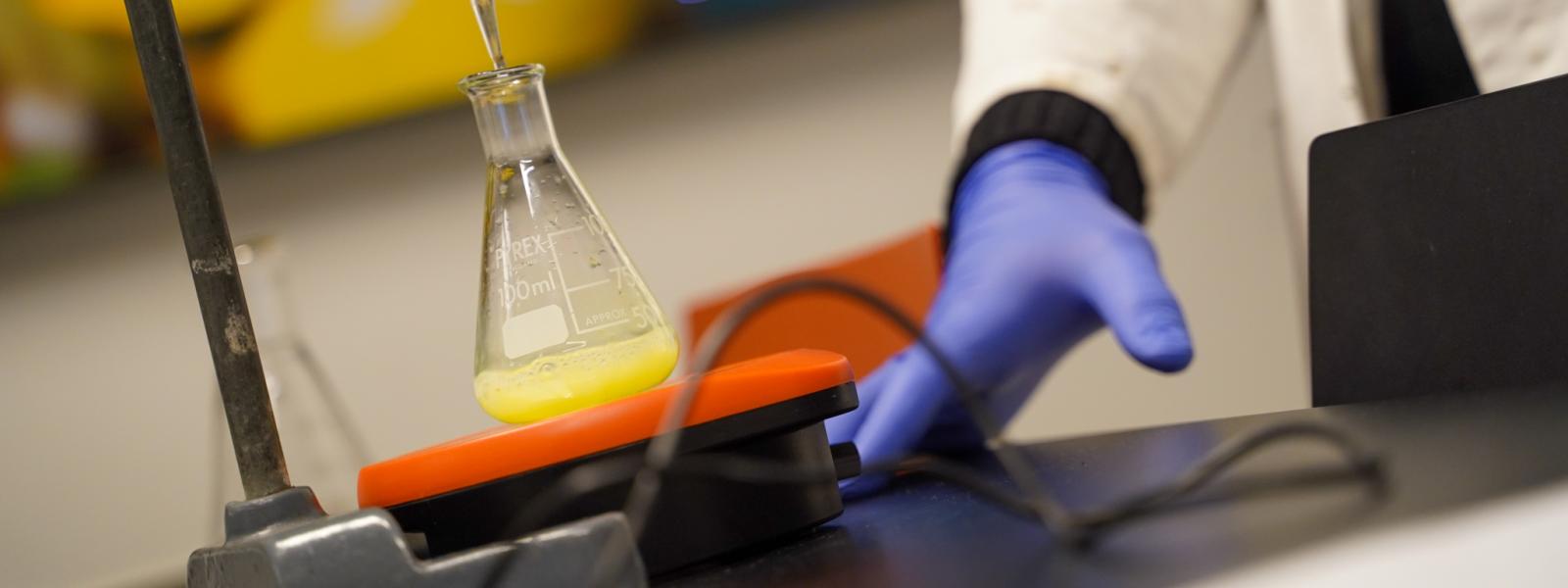 Et par hænder med blå gummihandsker er i gang med at lave kemiforsøg med gul væske i et glas.