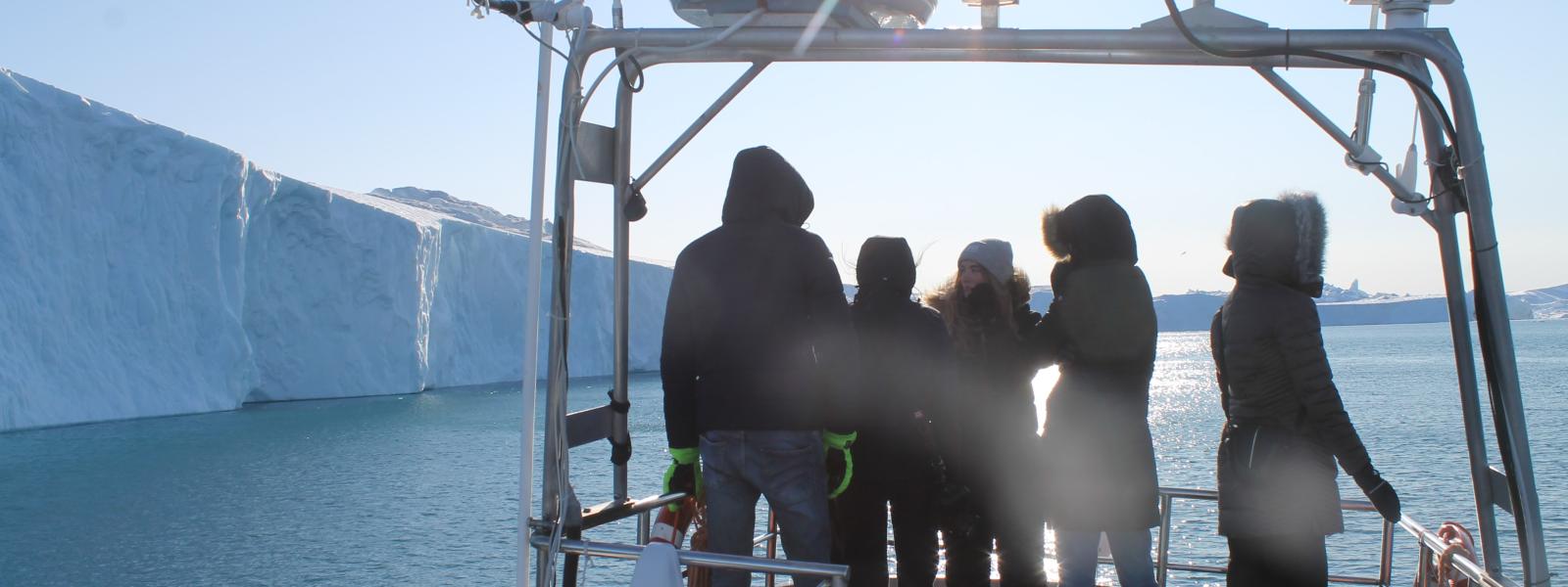 Fem elever står på en båd, der sejler i en fjord. Der er store isbjerge til venstre for dem.